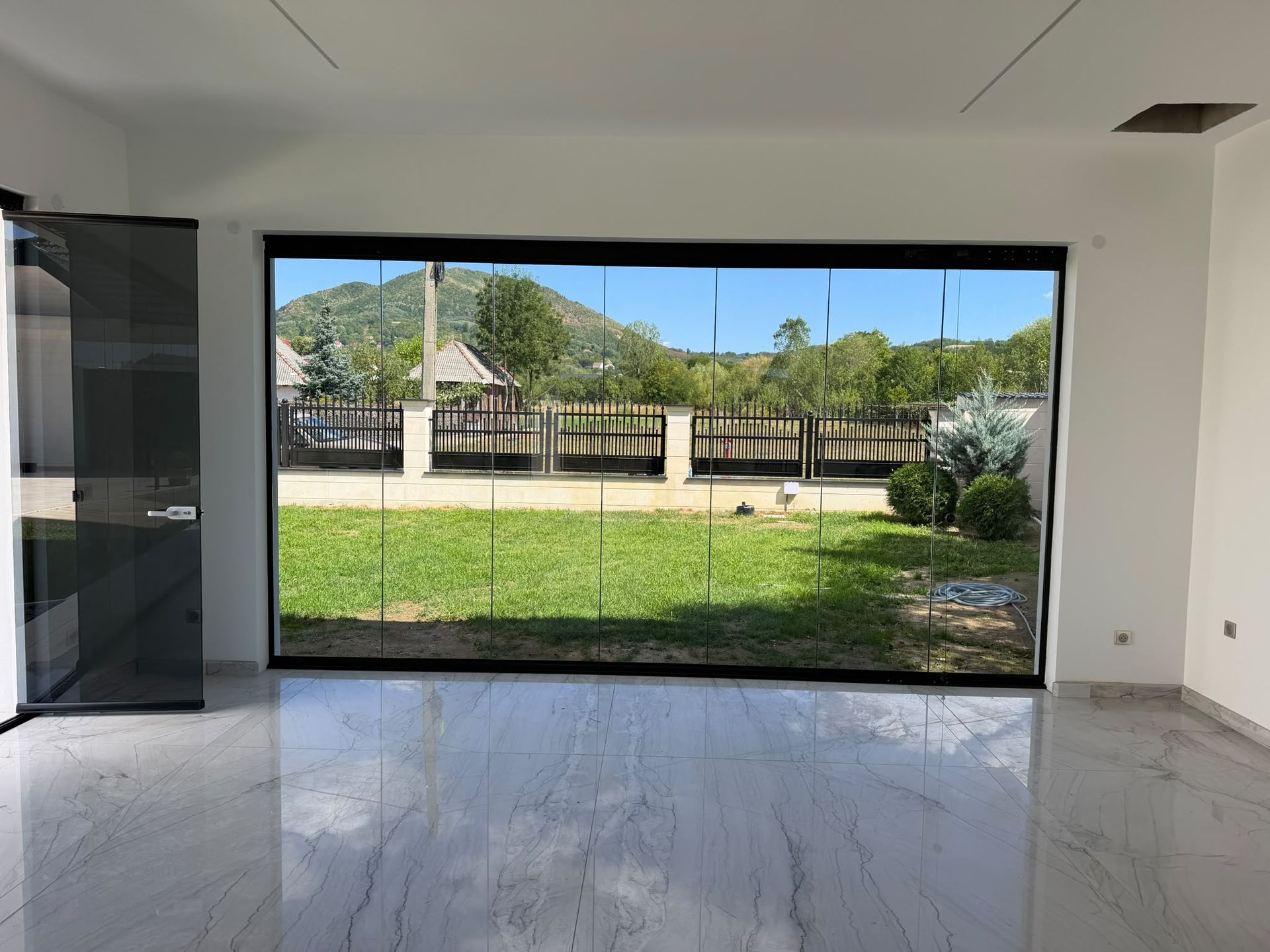Vedere din interiorul terasei prin sticla Valedo BKS-T spre gazon, gardul alb și dealul din fundal, județul Satu Mare