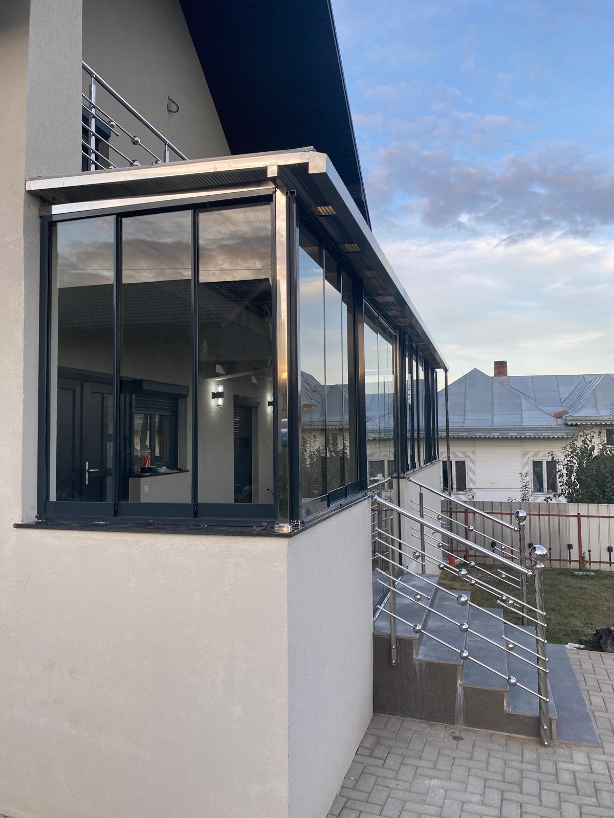Colțul vitrat al terasei la amurg, cu lumina aprinsă înăuntru și panourile de sticlă care preiau culoarea cerului