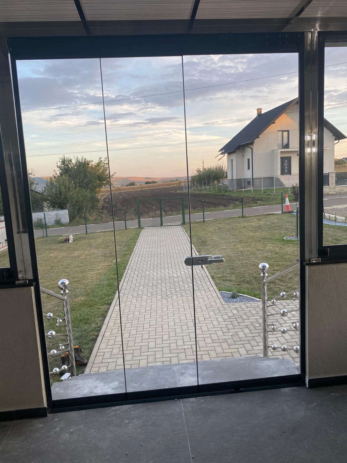 Vedere dinspre interiorul terasei spre curte, prin panoul de sticlă glisat la apus, cu profilul de aluminiu antracit în prim-plan